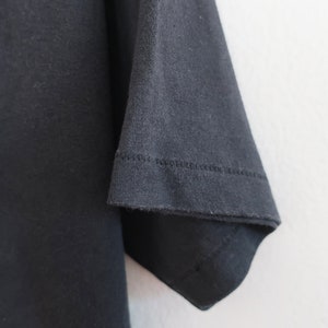 Op de afbeelding: Zwart t-shirt met een kleurrijke afbeelding van een boslandschap. De mouw is zichtbaar, met de zoom en de textuur van de stof. Het ontwerp bevat groene bomen en een blauwe lucht.