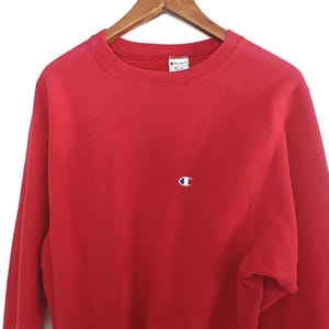 Peut inclure: Un sweat-shirt Champion rouge avec un col rond et des manches longues. Le sweat-shirt présente un petit logo blanc et bleu sur la poitrine. Le vêtement est suspendu à un cintre en bois sur fond blanc.