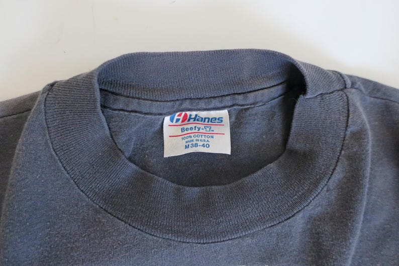 May include: A gray Hanes Beefy-T t-shirt tag with white text that reads "Hanes Beefy-T", "100% Cotton", "Made in U.S.A.", and "M 38-40".