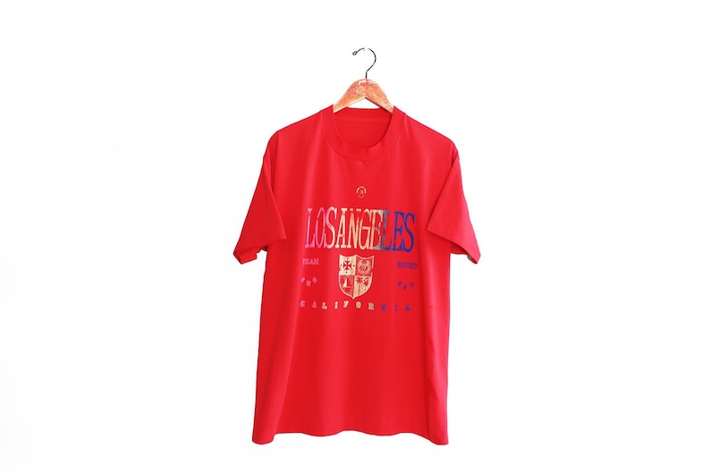 May include: Red short-sleeved t-shirt with a graphic print of "LOS ANGELES" in rainbow colors with a crest and "CALIFORNIA" underneath.