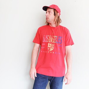 May include: A red t-shirt with a graphic print of a crest and the text "LOS ANGELES" and "CALIFORNIA". The person is wearing a red baseball cap.