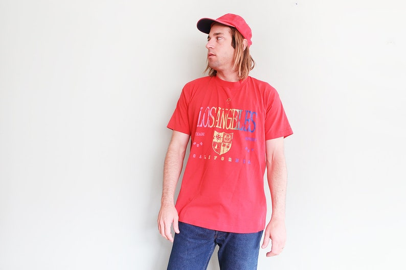 May include: A red t-shirt with a graphic print of a crest and the text "LOS ANGELES" and "CALIFORNIA". The person is wearing a red baseball cap.