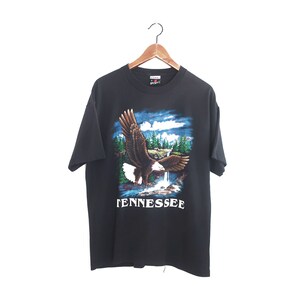 Op de afbeelding: Zwart T-shirt met een afbeelding van een adelaar die boven een waterval en bomen vliegt. De tekst "TENNESSEE" staat in het wit gedrukt. Het T-shirt hangt aan een houten hanger.