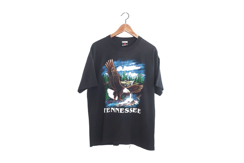Op de afbeelding: Zwart T-shirt met een afbeelding van een adelaar die boven een waterval en bomen vliegt. De tekst "TENNESSEE" staat in het wit gedrukt. Het T-shirt hangt aan een houten hanger.