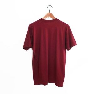 Op de afbeelding: Een effen maron T-shirt hangend aan een houten hanger tegen een witte achtergrond.