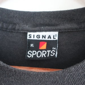Op de afbeelding: Close-up van een zwarte t-shirtkraag met een label. Op het label staat "SIGNAL SPORTS" in witte tekst, met een rood en oranje logo. Het shirt is maat XL.