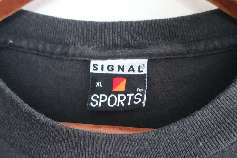 Op de afbeelding: Close-up van een zwarte t-shirtkraag met een label. Op het label staat "SIGNAL SPORTS" in witte tekst, met een rood en oranje logo. Het shirt is maat XL.