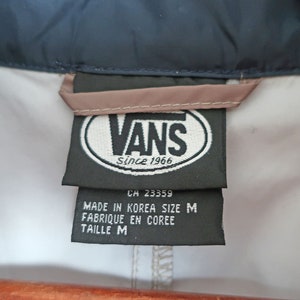 Pu&ograve; includere: Un'etichetta del marchio Vans in bianco e nero con il testo "Vans Since 1966" e "CH 23359" in un ovale bianco. Sotto l'ovale si trova il testo "Made in Korea Size M Fabrique en Coree Taille M".