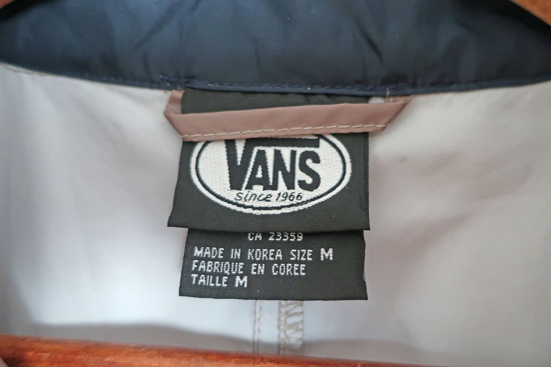Pu&ograve; includere: Un'etichetta del marchio Vans in bianco e nero con il testo "Vans Since 1966" e "CH 23359" in un ovale bianco. Sotto l'ovale si trova il testo "Made in Korea Size M Fabrique en Coree Taille M".