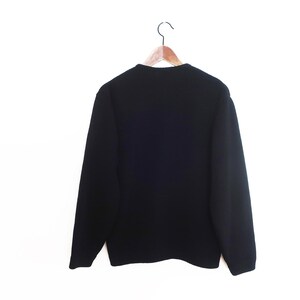 May include: A black sweater hangs on a wooden hanger against a white background. The sweater has long sleeves and a crew neck. The back of the sweater is visible, showing the full shape and design.