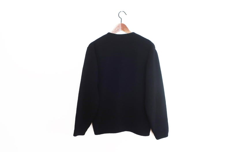 May include: A black sweater hangs on a wooden hanger against a white background. The sweater has long sleeves and a crew neck. The back of the sweater is visible, showing the full shape and design.
