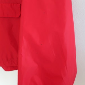 May include: A red jacket with a zip and a flap pocket on the front. The jacket has a few small black marks on the sleeve.