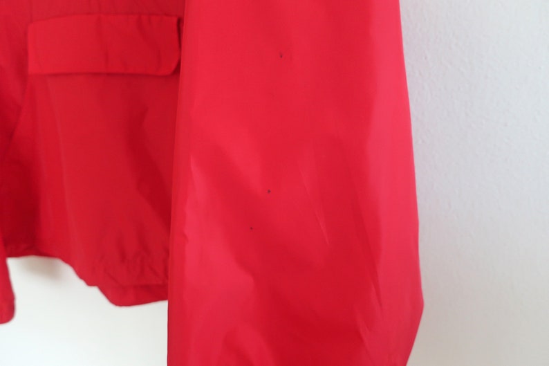 May include: A red jacket with a zip and a flap pocket on the front. The jacket has a few small black marks on the sleeve.