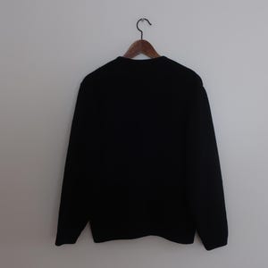 May include: A black crew neck sweater hangs on a wooden hanger against a white wall. The sweater has long sleeves and a simple, classic design. The garment is made of a soft, likely knitted material.