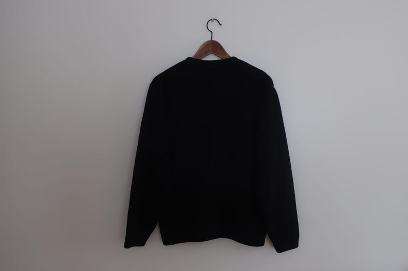 May include: A black crew neck sweater hangs on a wooden hanger against a white wall. The sweater has long sleeves and a simple, classic design. The garment is made of a soft, likely knitted material.