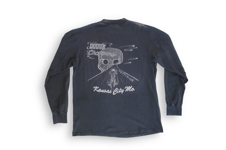 May include: A dark blue long-sleeved t-shirt with a white graphic of a skull riding a motorcycle on a road with the text "DOOGS Choppers" and "Kansas City Mo."