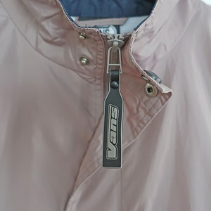 Pu&ograve; includere: Una giacca rosa con cerniera e un tiretto della cerniera nero con la scritta "Vans" in lettere bianche.