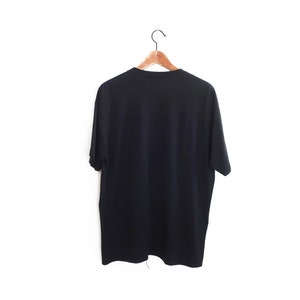 Op de afbeelding: Een effen zwart t-shirt hangt aan een houten kleerhanger tegen een witte achtergrond. Het shirt met korte mouwen is gemaakt van een zacht materiaal en wordt van achteren getoond. De hanger is bevestigd aan een metalen haak.