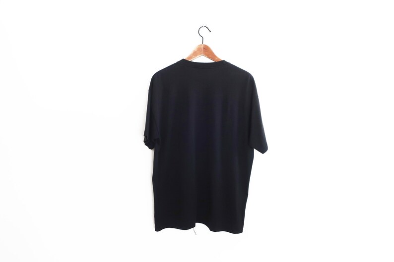 Op de afbeelding: Een effen zwart t-shirt hangt aan een houten kleerhanger tegen een witte achtergrond. Het shirt met korte mouwen is gemaakt van een zacht materiaal en wordt van achteren getoond. De hanger is bevestigd aan een metalen haak.