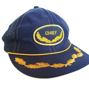 May include: A blue and gold trucker hat with the word "CHIEF" embroidered on the front. The hat has a mesh back and a yellow trim.