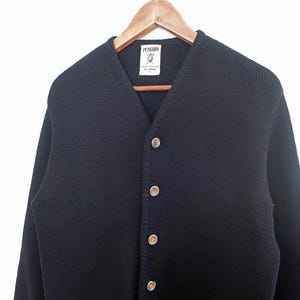 May include: A navy blue cardigan sweater with a V-neck and button closure. The sweater is hanging on a wooden hanger against a white background. The brand label "Penguin" is visible at the neckline.