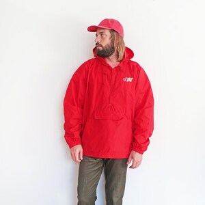 May include: A red hooded anorak jacket with a zip closure and a kangaroo pocket. The jacket features a colourful "GO-NY" logo on the front. The model is wearing a pink baseball cap and brown trousers.