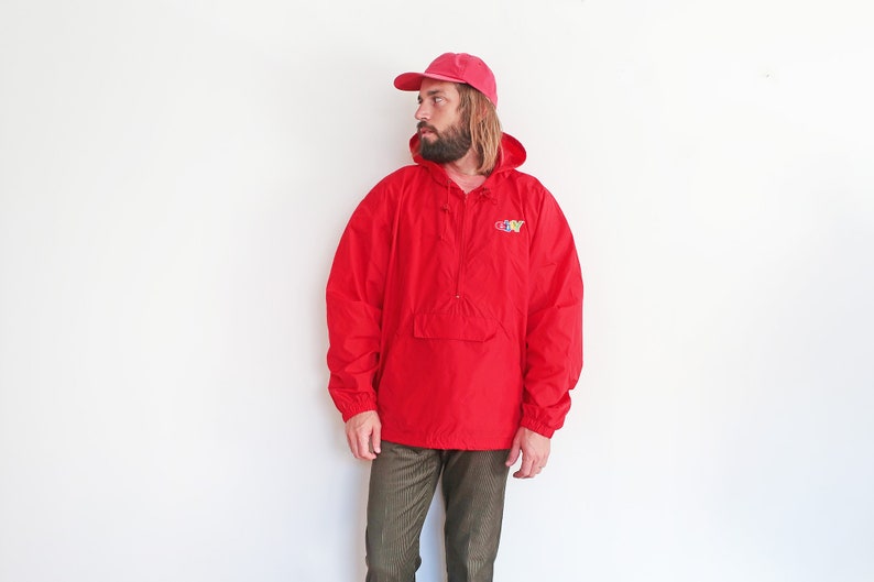 May include: A red hooded anorak jacket with a zip closure and a kangaroo pocket. The jacket features a colourful "GO-NY" logo on the front. The model is wearing a pink baseball cap and brown trousers.
