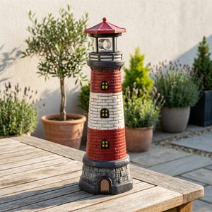 Pode incluir: Uma figura decorativa de farol com telhado vermelho, corpo cilíndrico com seções de tijolos vermelhos e brancos alternados, três janelas pequenas e uma base de pedra cinzenta com uma porta em arco na parte inferior.