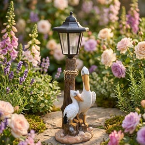 May include: A decorative solar-powered garden lamp with a black lantern top and a wooden post. Two white pelican statues are at the base of the lamp. The lamp is surrounded by colorful flowers and greenery.