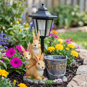 May include: A garden statue featuring three brown rabbits stacked together, with a black solar-powered lantern and a metal bucket that says "WELCOME". The statue is surrounded by colorful flowers.