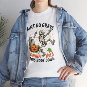 May include: A white t-shirt featuring a graphic of a dancing skeleton, a tombstone, and an orange jack-o'-lantern. The text reads AINT NO GRAVE GONNA HOLD THIS BODY DOWN in black and orange lettering. The shirt is worn under a blue denim jacket.
