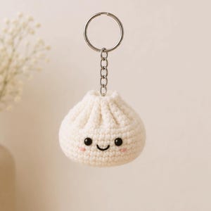 May include: A white, crocheted dumpling keychain with a smiling face, black eyes, and pink cheeks. The keychain has a silver ring and chain. The dumpling is a whimsical, handmade accessory.