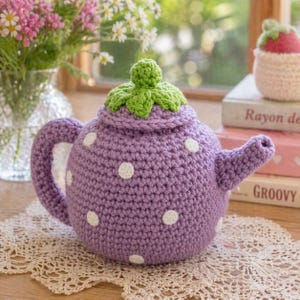 May include: A crocheted teapot in the shape of a strawberry. The teapot is purple with white polka dots and a green leaf-shaped lid. The handle and spout are also purple. The teapot is sitting on a lace doily.