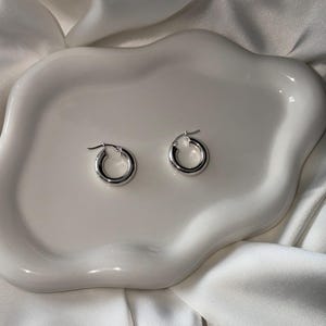 May include: Two silver-colored hoop earrings. The earrings are round and have a polished finish. They are displayed on a white, organically shaped ceramic dish, set against a backdrop of white fabric.