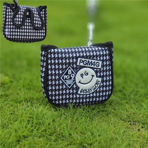 May include: A black and white houndstooth golf putter cover with the text "PGM4G", a smiling face, and the words "Ready Golf". The cover is on a green grassy surface.