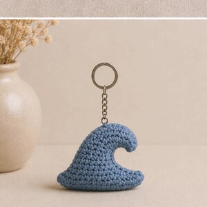 May include: A blue crocheted wave keychain with a silver-colored chain and key ring. The wave is a light blue color and has a textured appearance. The keychain is a handmade item.