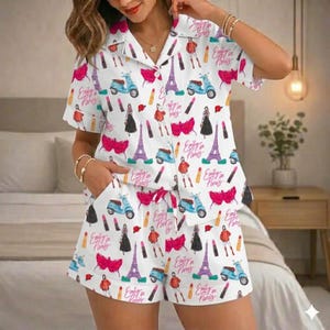 Puede incluir: Conjunto de pijama blanco con un estampado repetido de la Torre Eiffel, scooters, pintalabios e ilustraciones de moda. Tambi&eacute;n aparece el texto "Emily in Paris". El conjunto incluye una camisa con botones y pantalones cortos.