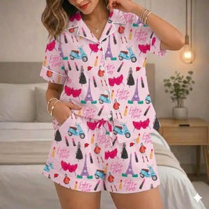 Puede incluir: Conjunto de pijama rosa claro con un estampado repetido de scooters, pintalabios, la Torre Eiffel e ilustraciones de moda. Tambi&eacute;n aparece el texto "Emily in Paris". El conjunto incluye una camisa abotonada con ribete blanco y pantalones cortos con cord&oacute;n.