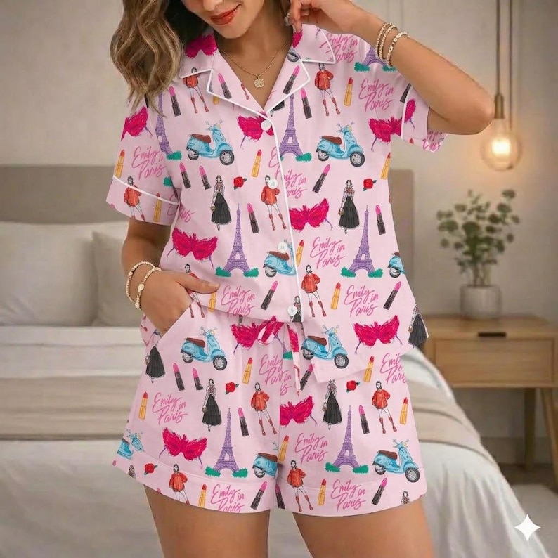 Puede incluir: Conjunto de pijama rosa claro con un estampado repetido de scooters, pintalabios, la Torre Eiffel e ilustraciones de moda. Tambi&eacute;n aparece el texto "Emily in Paris". El conjunto incluye una camisa abotonada con ribete blanco y pantalones cortos con cord&oacute;n.