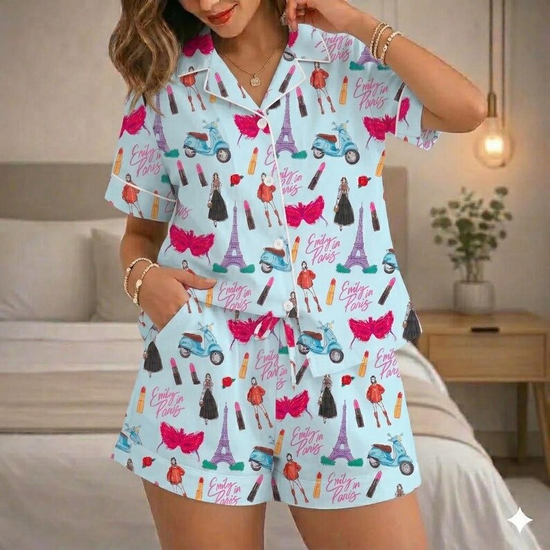 Puede incluir: Conjunto de pijama azul claro con un estampado repetido de la Torre Eiffel, scooters, pintalabios e ilustraciones de moda. El conjunto incluye una camisa de manga corta con botones y pantalones cortos a juego. El texto "Emily in Paris" tambi&eacute;n es visible.