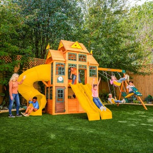 May include: A wooden playset with a yellow slide, swings, and a climbing structure. Children are playing on the slide and swings. The playset is on a green lawn, surrounded by trees.
