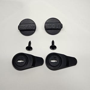 May include: A set of black hardware components, including two circular pieces with textured grips, two screws, and two lever-style pieces with keyhole-shaped openings. The items are arranged on a white surface.