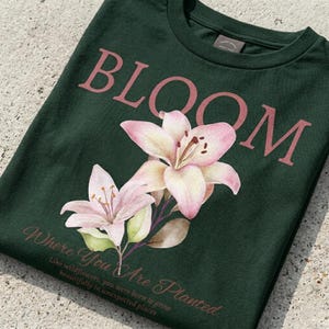 Puede incluir: Camiseta verde oscuro con la palabra "BLOOM" en color rosa empolvado sobre una ilustración de acuarela de lirios rosas y crema. Debajo de las flores, el texto dice "Where You Are Planted".
