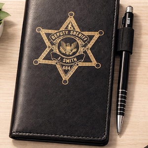May include: Black leather Deputy Sheriff badge notebook with a gold emblem. The emblem features a six-pointed star with the words "Deputy Sheriff" and "J. Smith 684". A black pen is attached to the side.