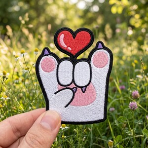 Op de afbeelding: Een geborduurde patch met een witte cartoon kattenpoot met roze kussentjes en paarse nagels, die een rood hart met een witte glans vasthoudt. Het ontwerp is omlijnd met dik zwart garen, wat zorgt voor een opvallende grafische uitstraling.