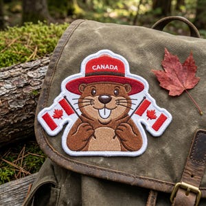 May include: An embroidered patch featuring a brown beaver wearing a red ranger hat with the word CANADA printed in white. The beaver is holding two small Canadian flags, one in each paw, with red and white stripes and a red maple leaf in the center.