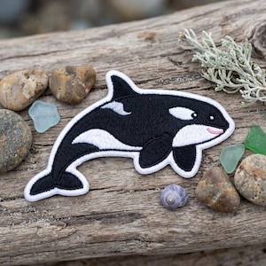 May include: An embroidered iron-on patch shaped like an orca whale. The patch features a black body with white underbelly, eye patch, and dorsal fin markings, outlined in a thick white border. The orca has a small pink stitched mouth.