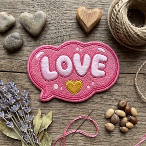 Op de afbeelding: Een roze geborduurde patch in de vorm van een tekstballon met het woord LOVE in witte letters en een klein goudkleurig hartje eronder. De patch heeft een donkerroze rand en is omringd door decoratieve items zoals stenen en lavendel.