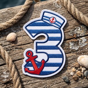 May include: An embroidered patch in the shape of the number three, featuring horizontal blue and white stripes. The top of the number is topped with a sailor hat with a small red anchor, and a large red anchor is positioned at the bottom curve.