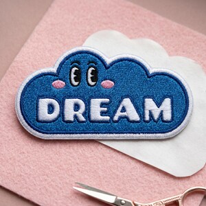May include: A blue cloud-shaped embroidered patch featuring the white text DREAM, two black and white cartoon eyes, and two small pink cheeks. The patch has a thick white border and is stitched with a dense thread texture throughout the design.